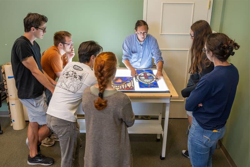 Professor Stephen Hartley stands over a light table illuminating a stained glass drawing while surrounded by students.