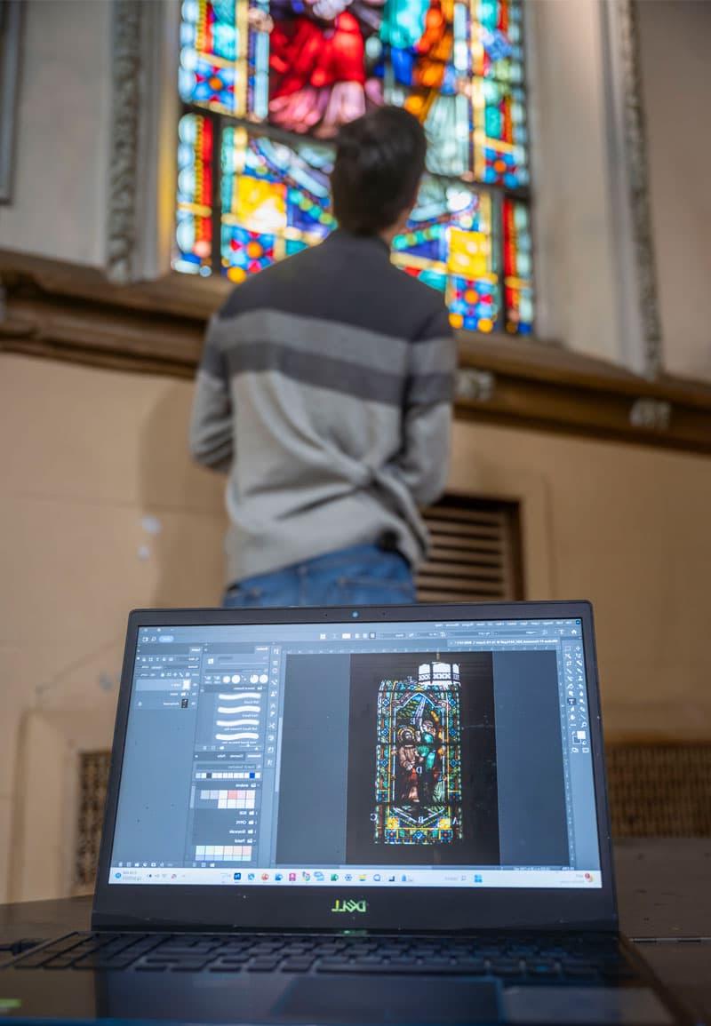 前景是一台带有彩色玻璃窗图像的笔记本电脑, 背景是一名男学生站在彩色玻璃窗前.